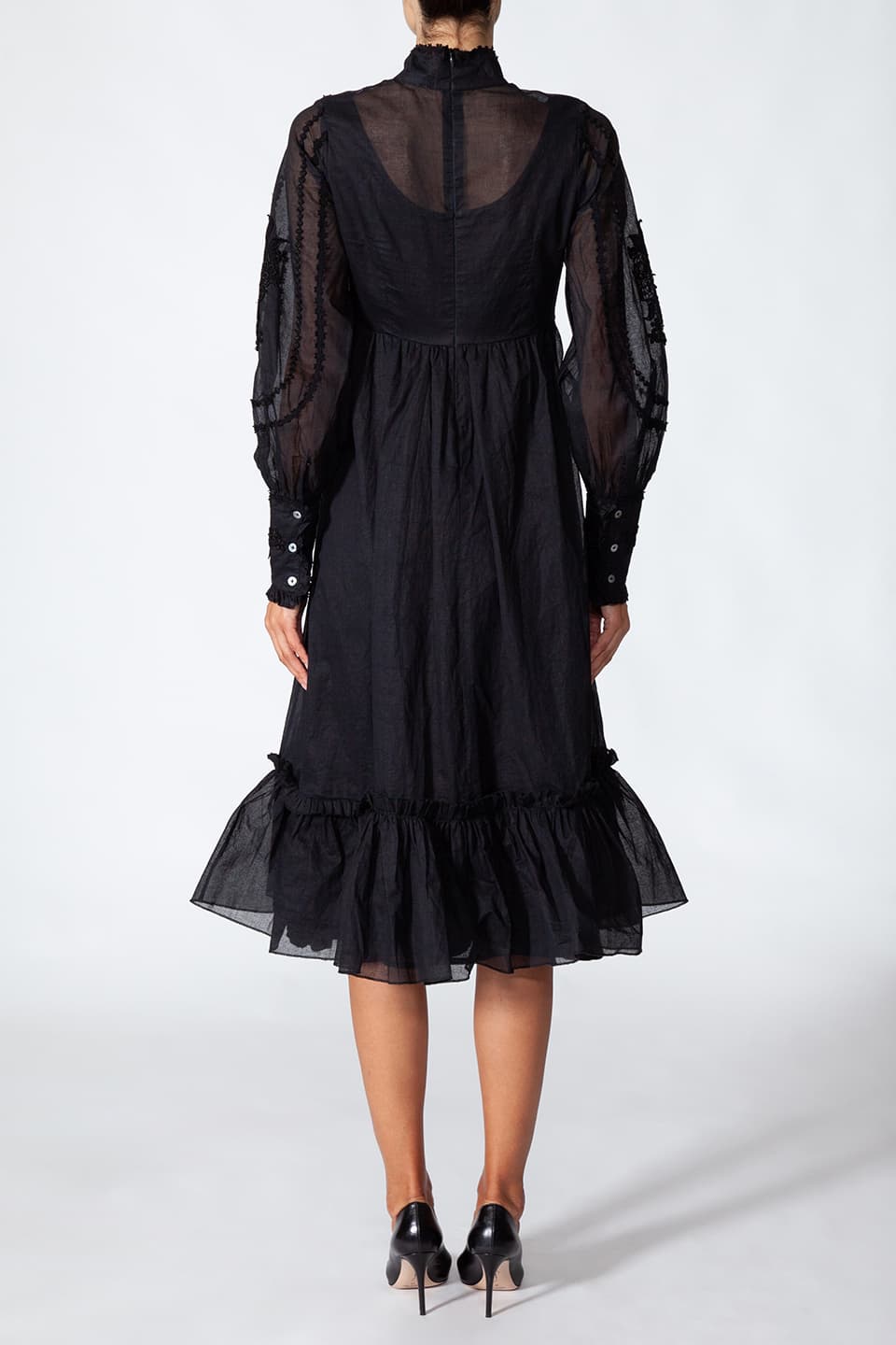 Model wearing long black dress in cotton. with floral embroidery on the chest, posing for back side view. Dress is from Fashion Designer Manoush collection.