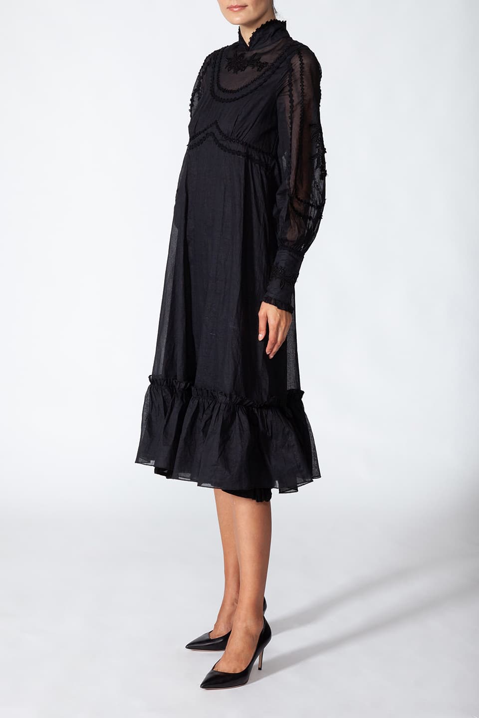 Model wearing long black dress in cotton. with floral embroidery on the chest, in pose from right side. Dress is from Fashion Designer Manoush collection.