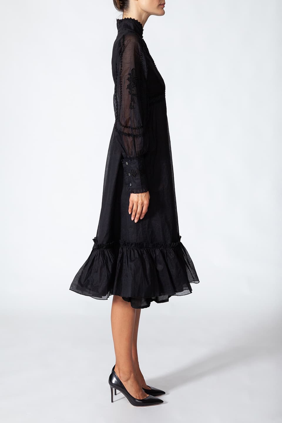 Model wearing long black dress in cotton. with floral embroidery on the chest, posing for left side view. Dress is from Fashion Designer Manoush collection.