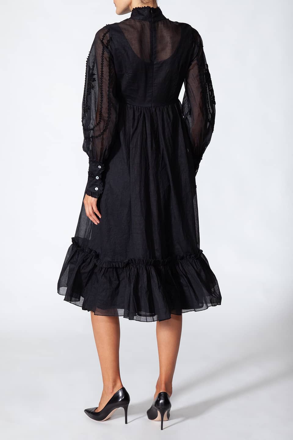 Model wearing long black dress in cotton. with floral embroidery on the chest, posing from behind. Dress is from Fashion Designer Manoush collection.